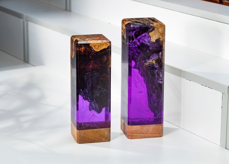 May include: Two rectangular decorative blocks. The top portion is a mix of brown and gold wood, while the lower portion is a vibrant purple resin. The blocks are set on a white surface.