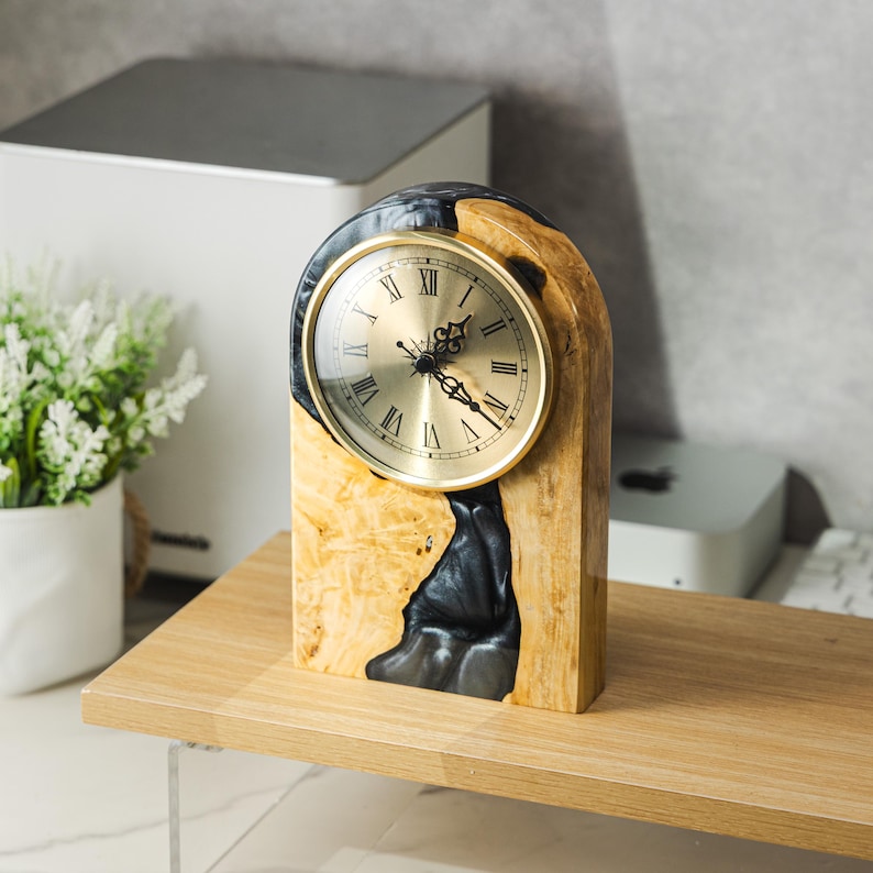 May include: A wooden desk clock with a gold-colored clock face and Roman numerals. The clock is encased in a light-colored wood with dark blue resin accents. The clock is sitting on a wooden surface.