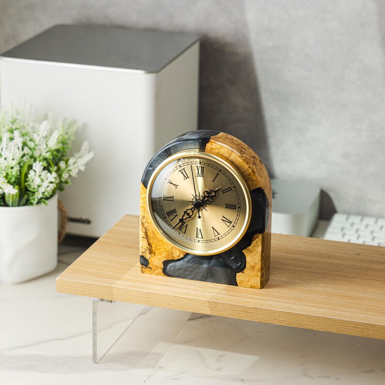 May include: A decorative desk clock with a gold-tone face and Roman numerals. The clock is encased in a unique wood and resin block, featuring a mix of natural wood grain and dark gray resin. The clock sits on a light wood shelf.