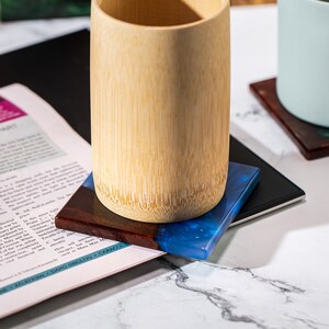 May include: A bamboo cup sits on a square coaster with a dark wood and blue resin design. The coaster rests on a black surface, with a magazine page visible beneath. Another coaster is in the background.