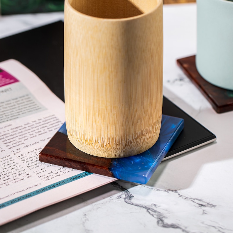 May include: A bamboo cup sits on a square coaster with a dark wood and blue resin design. The coaster rests on a black surface, with a magazine page visible beneath. Another coaster is in the background.