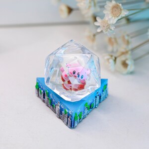 May include: A clear, faceted resin die with a pink and red cartoon character inside, set on a blue base with a cityscape design. The die is a unique decorative item.