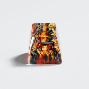 Op de afbeelding: Een heldere, rechthoekige keycap met een vurig ontwerp. De keycap heeft een mix van rood, oranje en zwart, die lijkt op een vulkanisch landschap. Een kleine, donkerblauwe structuur is zichtbaar.