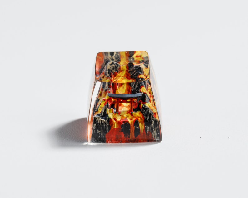 Op de afbeelding: Een heldere, rechthoekige keycap met een vurig ontwerp. De keycap heeft een mix van rood, oranje en zwart, die lijkt op een vulkanisch landschap. Een kleine, donkerblauwe structuur is zichtbaar.