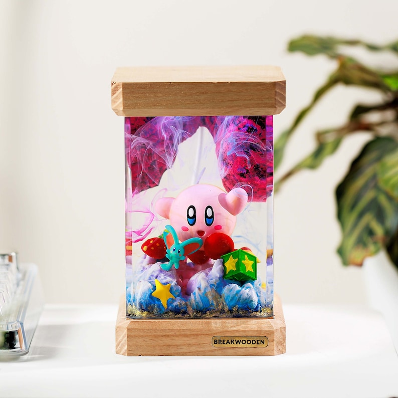 May include: A rectangular resin lamp with a wooden base and top, featuring a pink Kirby character figurine inside. The clear resin contains swirling purple and blue smoke-like patterns, a small green creature, red mushrooms, and yellow stars. The base has a metal plate labeled BREAKWOODEN.