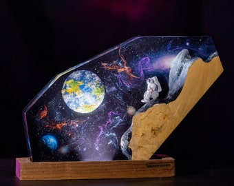 Può includere: Un'opera d'arte in resina che rappresenta una scena spaziale con un pianeta blu e verde, nebulose viola e blu e una piccola figura di astronauta su una superficie di legno. L'opera è intitolata "BreakWooden".