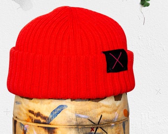 May include: A red knitted beanie with a black patch that has a pink 'X' on it. The beanie is sitting on top of a wooden barrel with colorful paint splatters.