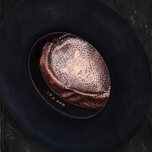 Peut inclure: Un chapeau noir à larges bords. L'intérieur présente un motif léopard avec le texte "LB & BAB" imprimé sur le bandeau intérieur. Le chapeau est un accessoire élégant.