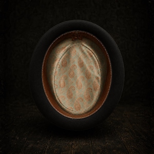 May include: A black felt hat viewed from the interior, featuring a tan leather sweatband and a light green satin lining decorated with a repeating copper-colored paisley pattern. The hat has a structured brim and a deep, rounded crown.