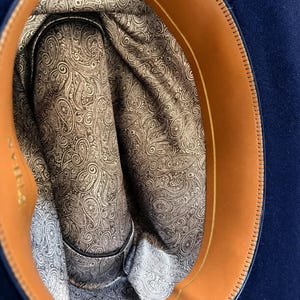 Pode incluir: O interior de um chapéu azul-escuro com uma faixa de suor de couro castanho e um forro de tecido com padrão paisley castanho e branco. O forro cobre a coroa e os lados, enquanto a faixa de couro contorna a borda interna do chapéu.