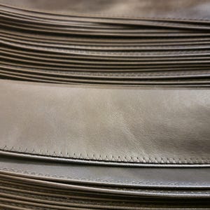 May include: A stack of multiple long, narrow strips of smooth, dark brown leather. Each individual strip features a clean, straight line of stitching running along one edge, creating a uniform and layered appearance across the entire collection.