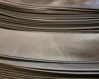 May include: A stack of multiple long, narrow strips of smooth, dark brown leather. Each individual strip features a clean, straight line of stitching running along one edge, creating a uniform and layered appearance across the entire collection.