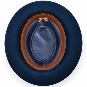 May include: A top-down view of a navy blue hat featuring a wide brim and a smooth, dark blue satin lining. The interior includes a tan leather sweatband and a small, centred tan ribbon bow detail positioned at the top of the inner crown.