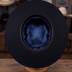 Peut inclure: Chapeau noir à large bord, vu de dessous. L'intérieur est doublé d'un tissu satiné bleu foncé. Le chapeau est présenté sur un présentoir en bois, soulignant sa forme et son design.