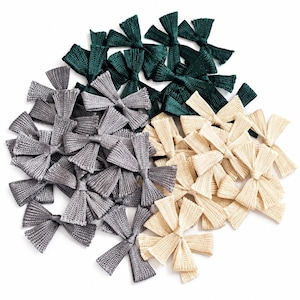 May include: A collection of small, textured fabric bows arranged in a cluster. The bows are colored in dark forest green, metallic silver-gray, and light cream, each featuring a ribbed surface texture and a central knot detail.