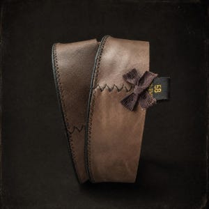 May include: A brown leather band featuring dark zigzag stitching along the edges. A small brown fabric bow is attached to the side, next to a black tag with the yellow text 58 and 7 1/4 printed on it. The band is curved into a loop shape.