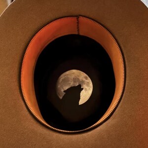 Peut inclure: Un chapeau beige encadre une pleine lune avec la silhouette d'un loup hurlant. L'intérieur du chapeau est doublé de tissu orange, créant une scène nocturne dramatique. Le bord du chapeau est visible.