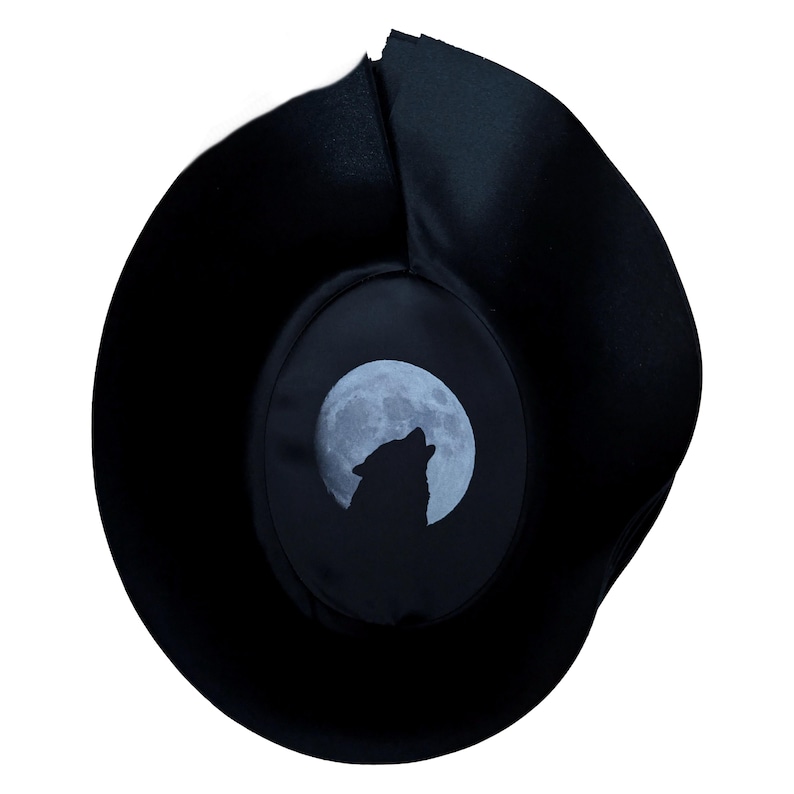 Pu&ograve; includere: Un cappello nero a tesa larga con una fodera interna circolare che mostra un'illustrazione grafica di un lupo che ulula alla luna piena. Il cappello ha una corona strutturata e una tesa curva e flessibile in tessuto scuro e liscio.