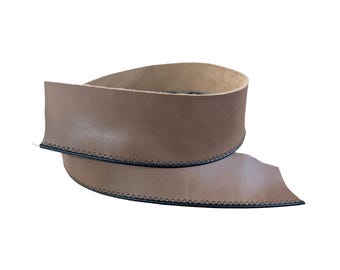 May include: A coiled strip of smooth, light brown leather featuring a dark black zigzag stitch pattern along both edges. The leather strip is curved into a circular shape, displaying a consistent tan colour on the surface and a visible texture.