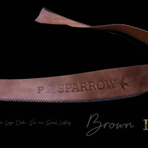以下が含まれることがあります： 「PA SPARROW」のエンボス加工の文字と鳥のロゴが入った茶色の革ストラップ。 ストラップはステッチされたエッジがあり、黒い背景に配置されています。 「Brown」の文字は筆記体で書かれています。
