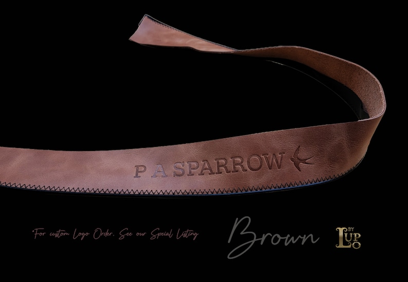 以下が含まれることがあります： 「PA SPARROW」のエンボス加工の文字と鳥のロゴが入った茶色の革ストラップ。 ストラップはステッチされたエッジがあり、黒い背景に配置されています。 「Brown」の文字は筆記体で書かれています。