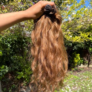 以下が含まれることがあります： ウェーブのかかった、明るい茶色のヘアエクステンションの束。緑の葉と青い空を背景に、手に持たれています。髪は自然なカールした質感で、暖かく赤みがかった茶色です。