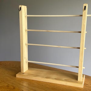 May include: A light-colored wooden display stand featuring a rectangular base and two vertical uprights connected by four horizontal dowel rods. Two small black metal hooks are attached to the top of each vertical wooden support post.