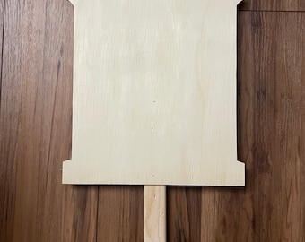 May include: A blank, unfinished wooden sign with a pointed stake for garden or yard display. The sign has a rectangular body with a decorative top and bottom edge. The wood has a light, natural color.