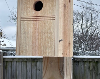 Könnte beinhalten: Ein Vogelhaus aus Holz mit einem runden Eingangsloch, montiert auf einem Holzpfosten. Das Vogelhaus hat ein schlichtes Design mit einem flachen Dach und einer strukturierten Holzoberfläche.