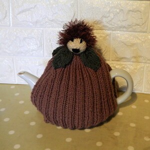 May include: A handmade, knitted tea cozy in a muted mauve color, shaped to fit a teapot. The cozy features a whimsical design with a face, dark green leaf accents, and a fluffy, dark brown hair-like element on top. The teapot is white.