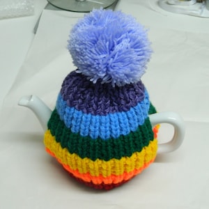 Peut inclure: Une théière en céramique blanche recouverte d'un cache-thé tricoté. Le cache-thé est rayé de couleurs arc-en-ciel : orange, jaune, vert, bleu et violet. Un gros pompon bleu clair et duveteux est posé sur le dessus.