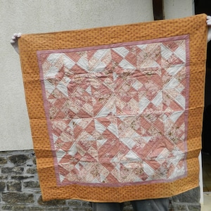 May include: A handmade quilt with a patchwork design. The quilt features a central pattern of pink and white triangles with floral prints, framed by a border of orange fabric with a small dot pattern. The quilt is held up, showcasing its details.