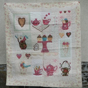 Puede incluir: Un edredón hecho a mano con fondo crema y borde floral. El edredón presenta imágenes aplicadas de teteras, tazas de té, cupcakes, corazones y arreglos florales en tonos rosa, marrón y verde. Un artículo decorativo encantador.