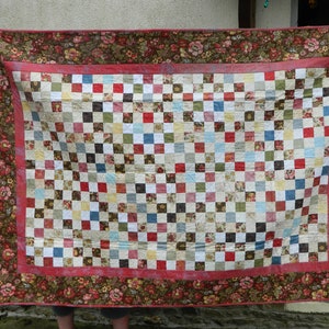 May include: A handmade quilt with a patchwork design. The center features a grid of small squares in various colors, including red, blue, and white. A floral border in shades of brown and red frames the quilt. The quilt is held up by unseen hands.