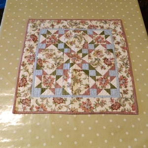 Peut inclure: Dessus de table carré matelassé avec un motif floral dans les tons crème, rouge, vert et bleu. Le centre présente un motif en étoile, et les bords sont bordés d'un imprimé floral assorti et d'une bordure rose.