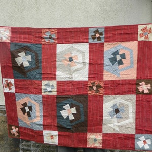 May include: A handmade quilt with a red background and a patchwork design. The quilt features alternating squares with geometric patterns in shades of brown, blue, pink, and white. The quilt is held up, showcasing its size and craftsmanship.