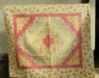 May include: A handmade quilt with a floral pattern in shades of yellow, pink, and green. The quilt features a central design surrounded by a pink border and a floral patterned edge. The quilt is likely made of cotton.
