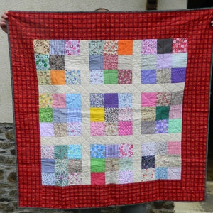 May include: A handmade patchwork quilt with a red plaid border. The quilt's center features a grid of colorful fabric squares in various patterns and colors, including floral, polka dot, and solid hues. The quilt is held up by unseen hands.