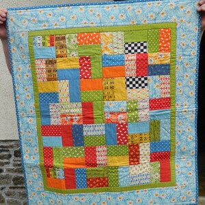 Può includere: Una colorata trapunta patchwork con un bordo azzurro a motivo di margherite. La trapunta presenta una varietà di quadrati di tessuto in arancione, verde, rosso, blu e giallo. Il design centrale include quadrati con numeri e motivi a scacchi.