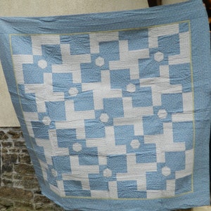 May include: A handmade quilt with a geometric pattern in light blue and white. The design features intersecting squares and octagons. The quilt has a light blue border with a floral pattern and a thin yellow trim.
