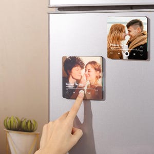 May include: Two square magnets with photos of couples, resembling music player interfaces. One magnet has a finger pressing a button. The text "Because You Live" is visible on the magnets. The magnets are attached to a silver refrigerator.