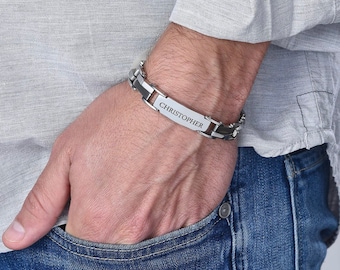 May include: A silver-toned bracelet with black accents and the name "CHRISTOPHER" engraved on a rectangular plate. The bracelet is worn on a wrist, with a light gray shirt and blue jeans in the background.
