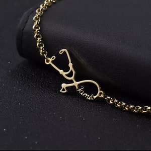 May include: Gold necklace featuring a stethoscope charm with the name "Jamie" engraved on it.