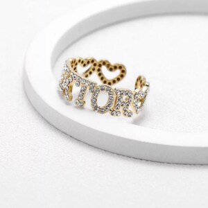 May include: Gold ring with the word "amore" spelled out in rhinestones. The ring is open and has two small hearts on the side.