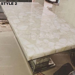 Puede incluir: Una mesa de tapa de ágata blanca con acabado pulido. La tapa de la mesa está hecha de muchas piezas pequeñas de ágata que se han unido. La tapa de la mesa está soportada por una base de metal plateado.