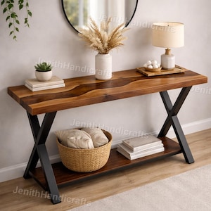 Op de afbeelding: Een houten console met een donkerbruin blad en een onderste plank. De tafel heeft zwarte metalen poten in een X-vorm. Boven de tafel hangt een ronde spiegel, die is versierd met een lamp, vaas en mand.
