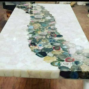 Puede incluir: Una mesa de madera blanca con una incrustación decorativa de ágata. El ágata es una variedad de calcedonia, una forma criptocristalina de sílice, y se caracteriza por su aspecto bandeado. El ágata está dispuesta en un patrón fluido, creando un diseño único y llamativo.
