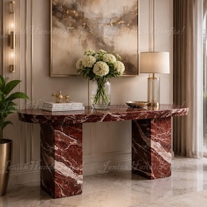 May include: A rectangular console table crafted from deep red marble with prominent white veining, featuring two thick, block-style legs. The table surface holds a clear glass vase filled with white hydrangea flowers, a small gold figurine, and a lamp.