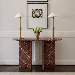 May include: A dark red marble console table with a smooth oval top and two vertical slab legs. Two tall brass lamps with white conical shades sit on the table, flanking a gold-toned vase filled with a bouquet of light green hydrangea flowers.