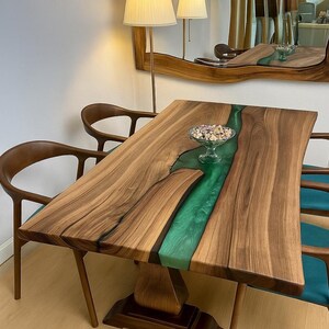 Puede incluir: Mesa de comedor de madera con un río de resina epoxi verde en el centro. La mesa tiene un acabado de vetas de madera natural y está sostenida por una base de pedestal. Se ven dos sillas de madera con cojines turquesa.